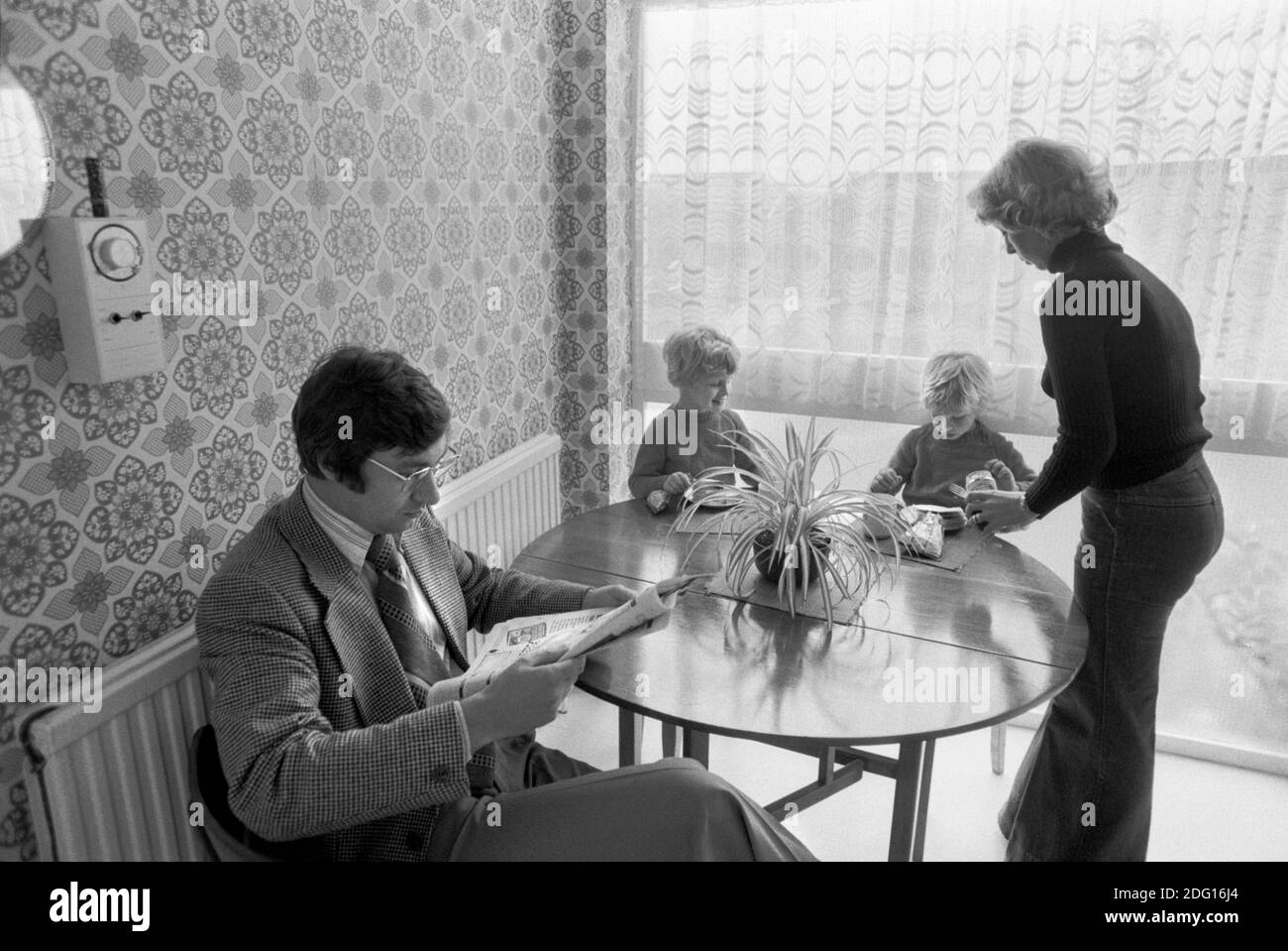1970s England, a middle income professional couple at home with their two children. Family life kitchen dinner  mother making afternoon tea, sandwiches for the two boys, and a cup of tea for her husband who is reading the newspaper and paying no attention at all or interest in his children. 1977 New town modern housing development Milton Keynes Buckinghamshire UK HOMER SYKES Stock Photo