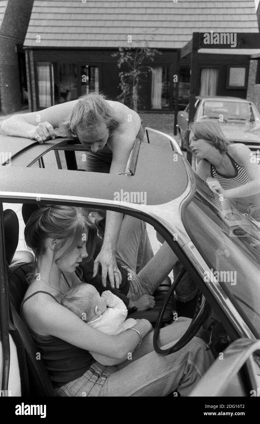 1970s professional middle class couple outside their new modern home on a new housing development. Father going to touch his new baby, mother looking on. Baby being held by girlfriend who is up for the day to see the new house and baby. Hanging out by the family car that is parked out side their house. 1977 HOMER SYKES Stock Photo