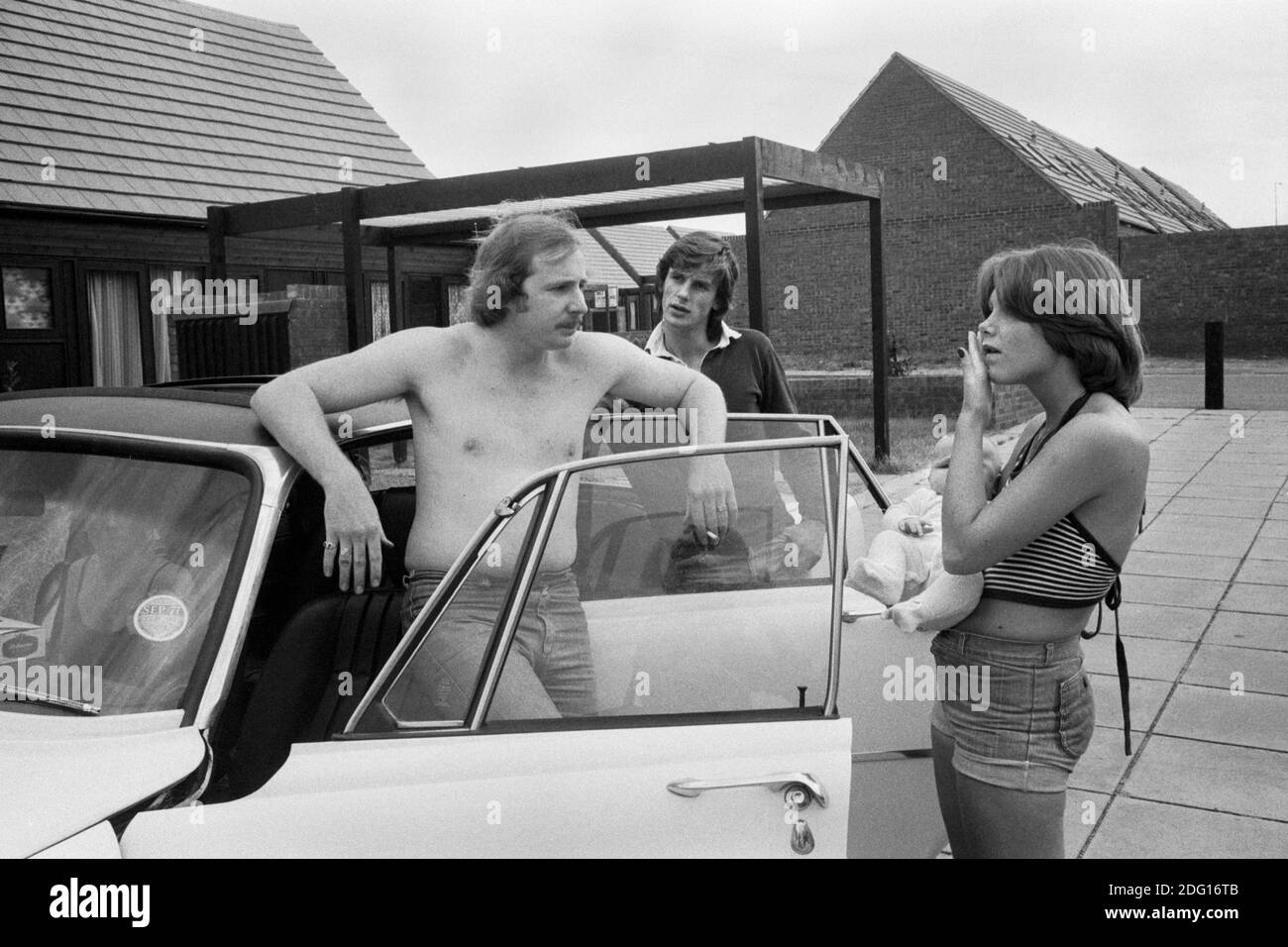 1970s UK, professional middle class couple outside their new modern home on a recently built new housing development. Hanging out with the new baby in arms by their car that is parked out side their home. The shirtless man is the car owner and father.   Its summer  and a friend is up for the day to see the new house. 1977 England. HOMER SYKES Stock Photo
