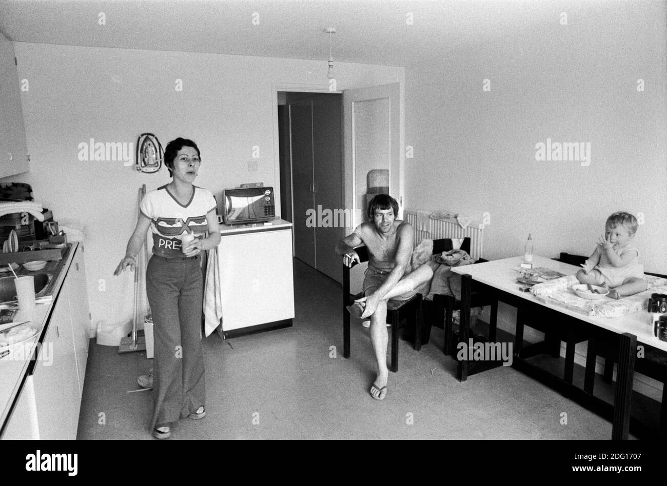 Modern living 1970s working class family life, mother, father and baby who is sitting on the kitchen table in the kitchen of their three bedroom home on a new modern housing development new town England 1977 Note the iron on the wall out of the way behind the woman's head. HOMER SYKES. Stock Photo