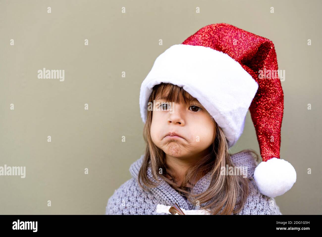 Cute little girl wearing Santa Claus hat against a green wall background outdoors in christmas time - Little girl sulking for Xmas gift - Funny face, Stock Photo
