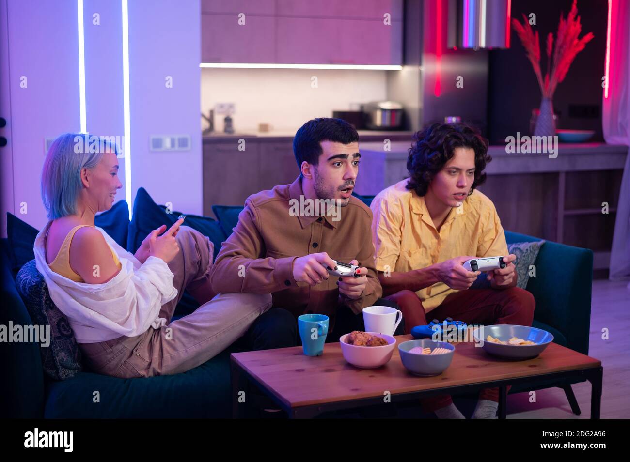 Two male friends excited to play video games at home while a girl sitting  with them on a couch and using mobile smart phone Stock Photo - Alamy