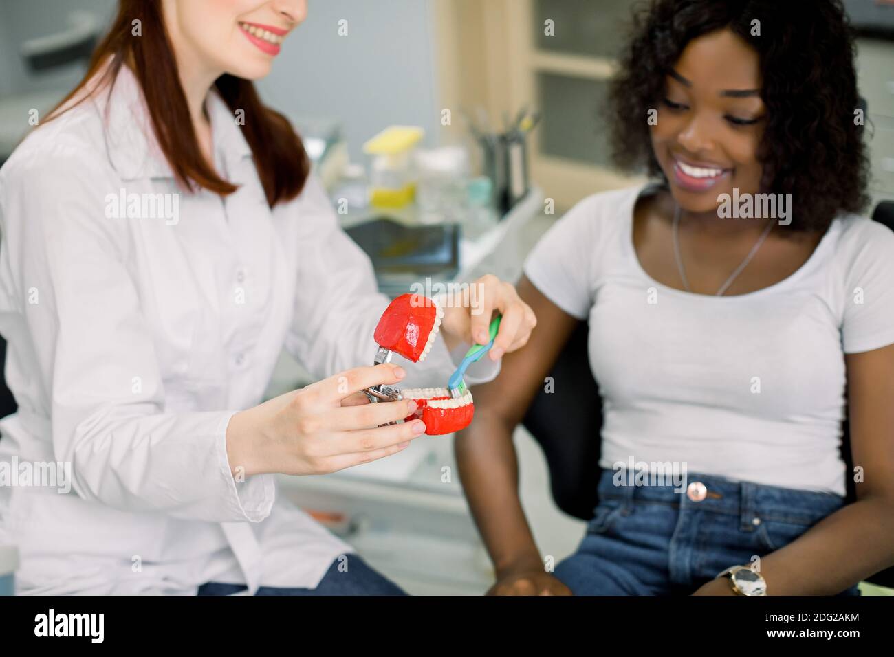 Healthy teeth, cleaning with toothbrush. Young smiling woman dentist explaining importance of oral hygiene on jaw model to smiling African female Stock Photo