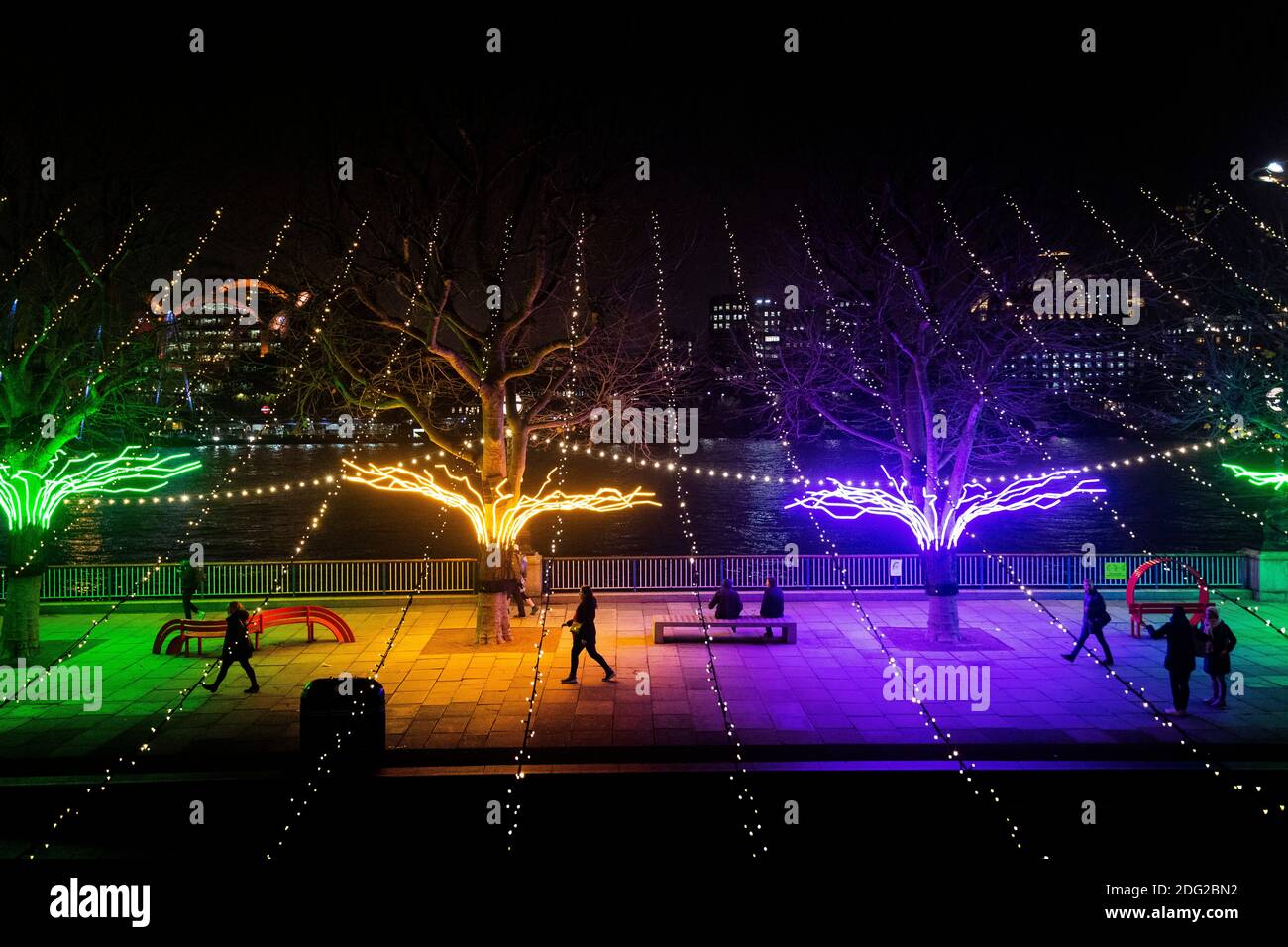 Pedestrians pass 'Lumen' by David Ogle, a row of riverside plane trees illuminated by neon flex, part of 'Winter Light', a new open-air exhibition at the Southbank Centre in London, which features over 15 artworks and new commissions that make use of light, colour and animation from a number of international artists. Stock Photo