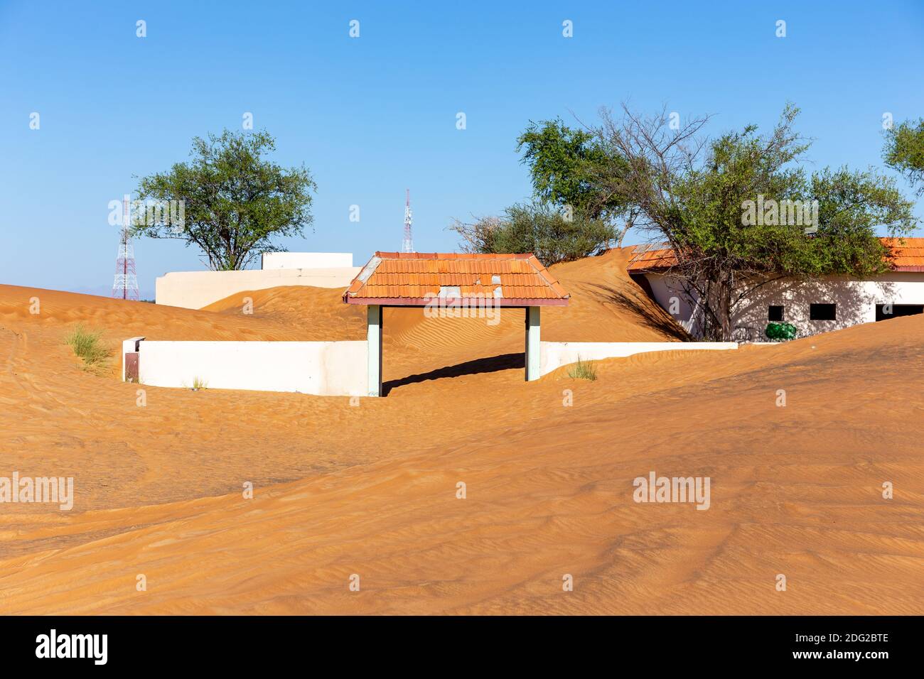 White entry gate buried in sand in Al Madam ghost village, with sand dunes and wild ghaf trees. Stock Photo