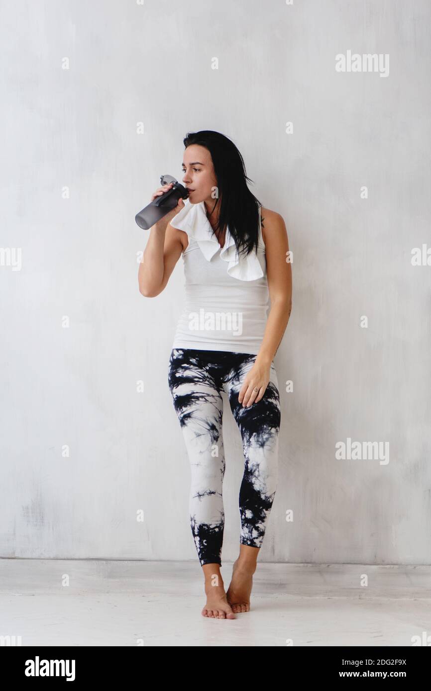 Fit young woman tired after training drinking water leaning on the white wall. Stock Photo