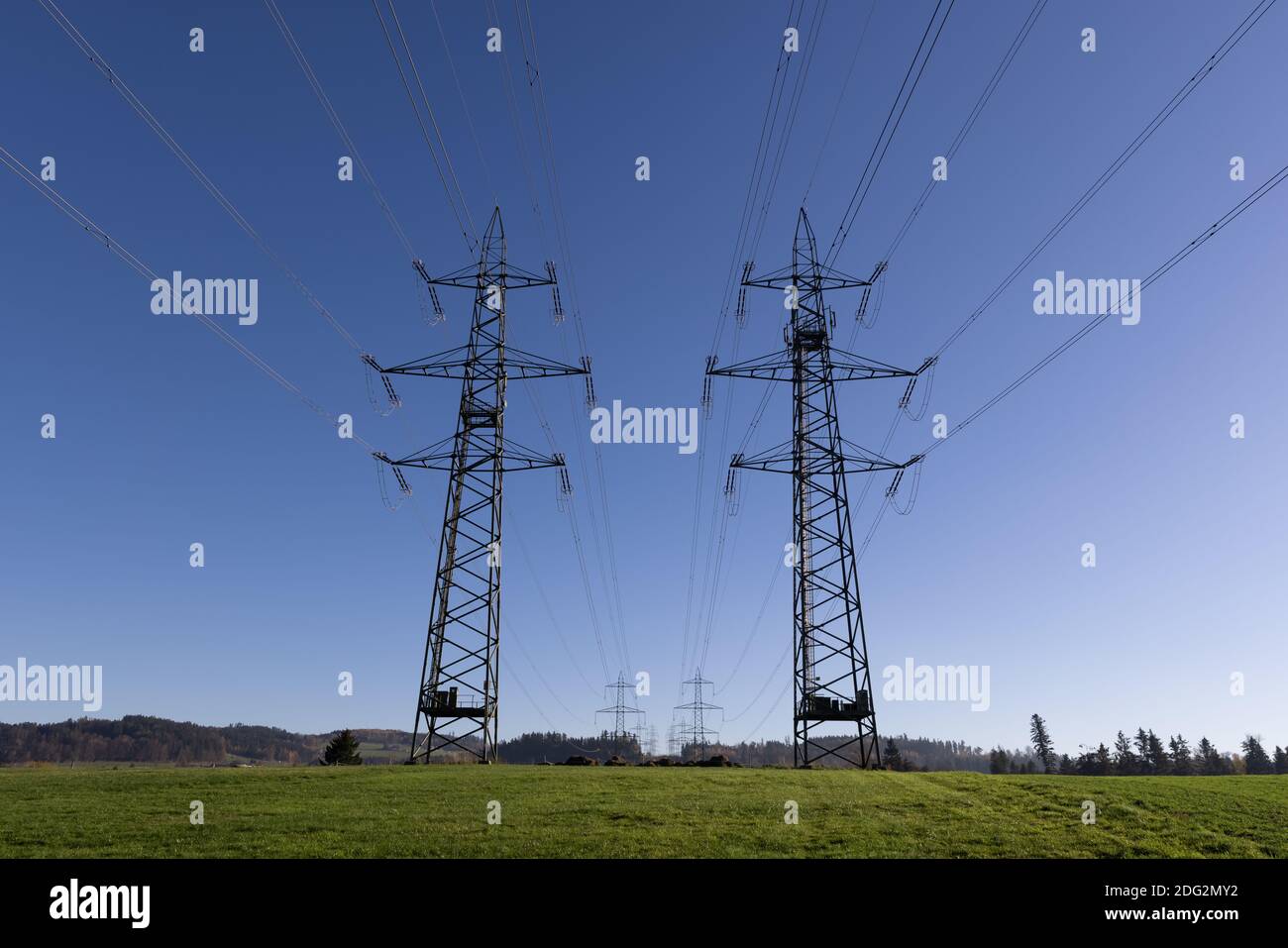 two large high voltage power poles in front of a bright blue sky on a green meadow in autumn, more power poles can be seen in the background, electric Stock Photo