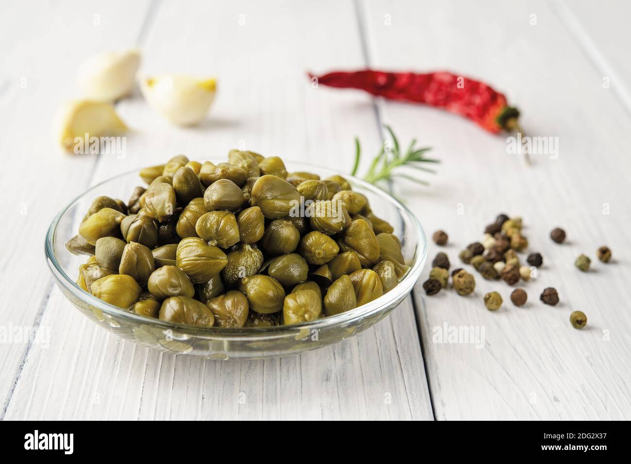 Pickled capers in a glass saucer, red hot pepper pod, garlic and black pepper grains on a white