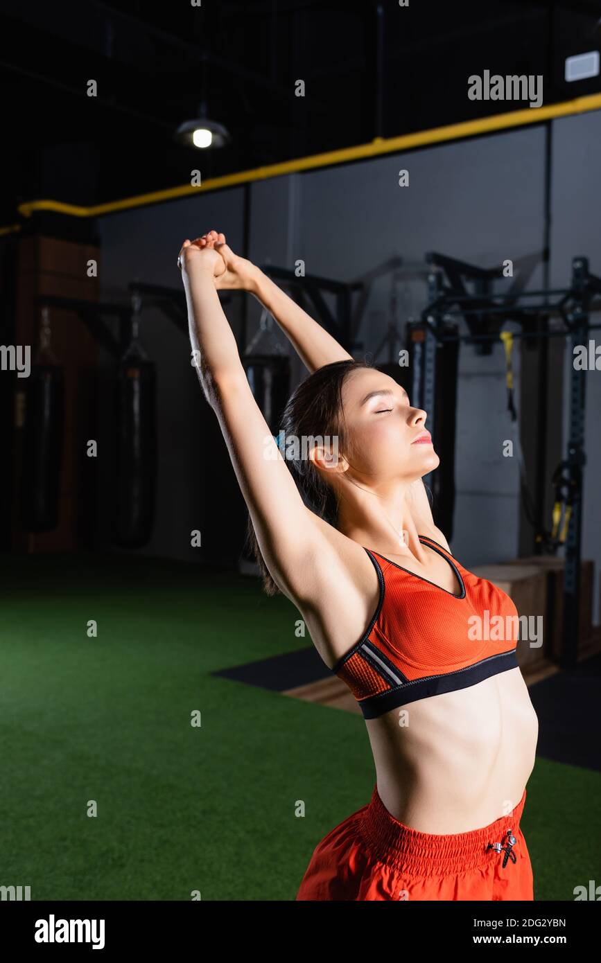 sportive woman in sports bra stretching hands with closed eyes in gym Stock Photo