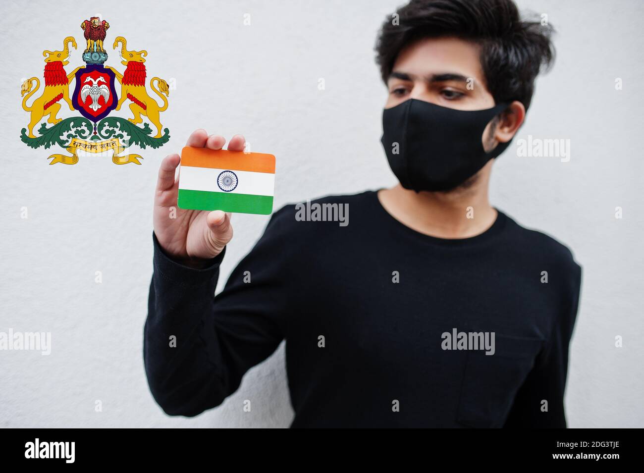 Indian man wear all black and face mask, hold India flag in hand isolated on white background with Karnataka state emblem . Coronavirus India states a Stock Photo