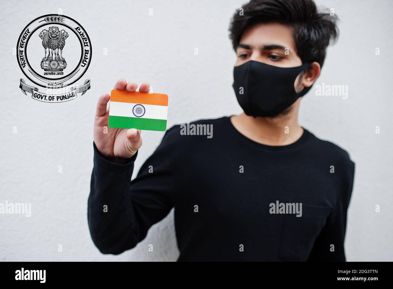 Indian man wear all black and face mask, hold India flag in hand isolated on white background with Punjab state emblem . Coronavirus India states and Stock Photo
