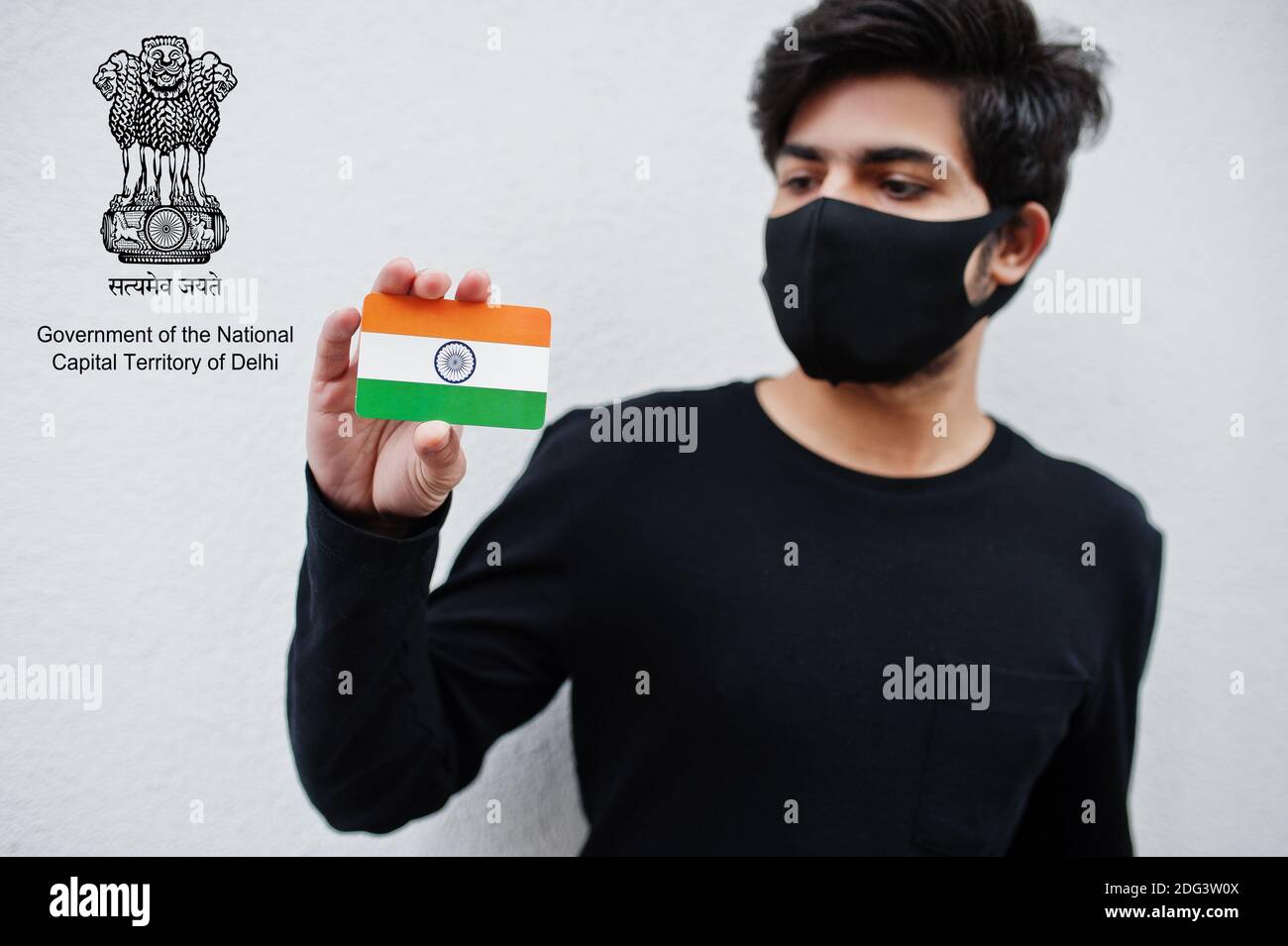 Indian man wear all black and face mask, hold India flag in hand isolated on white background with Delhi union territory emblem . Coronavirus India st Stock Photo