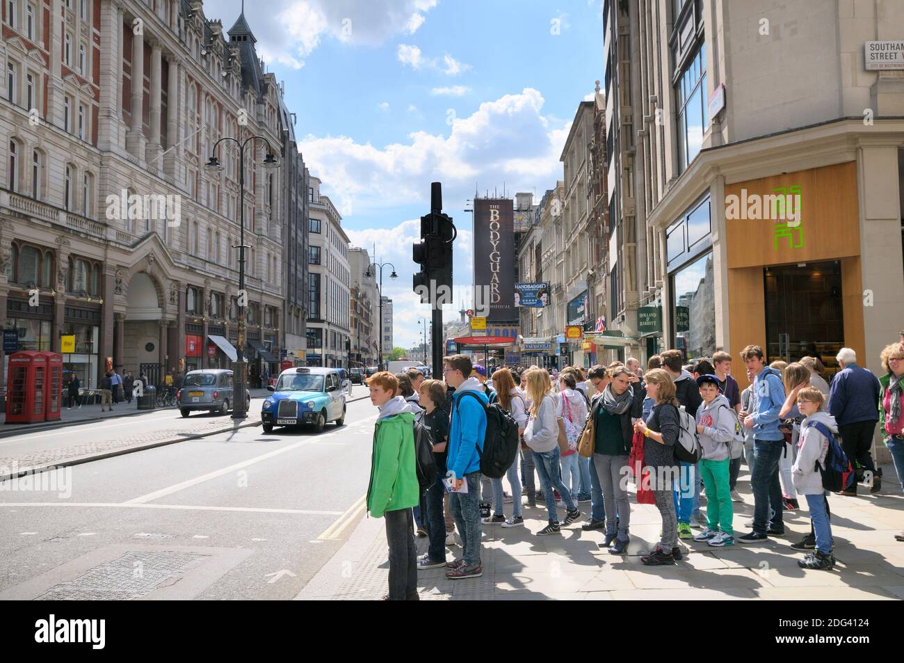A supervised group of young teenage European tourists waiting to cross the road at traffic lights on The Strand in central London, England, UK Stock Photo