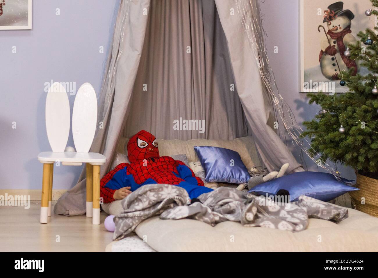 A tired child dressed in a Spiderman costume with mask is lying on a bed with canopy and pillows near a Christmas tree Stock Photo