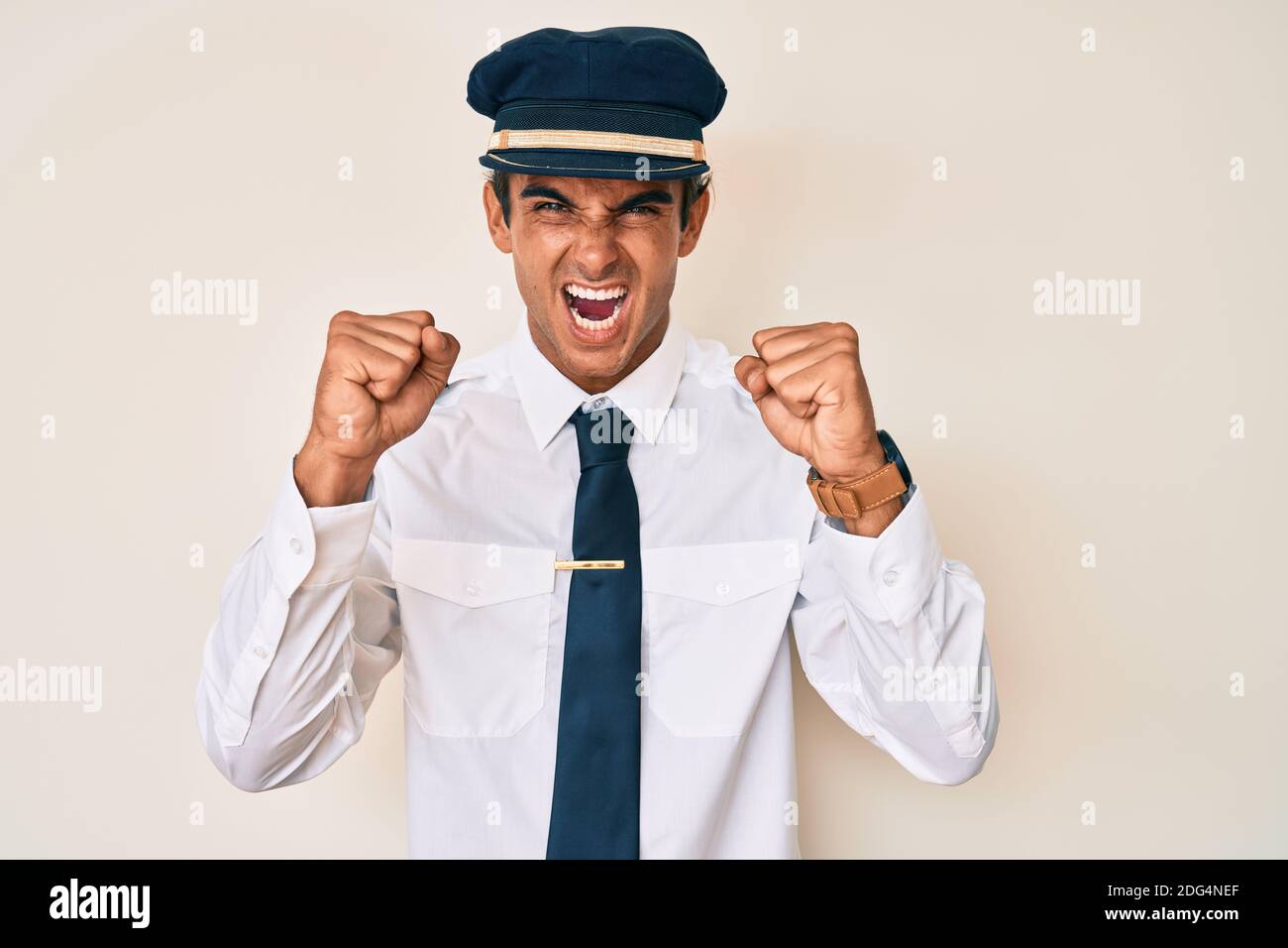 Young hispanic man wearing airplane pilot uniform angry and mad raising ...