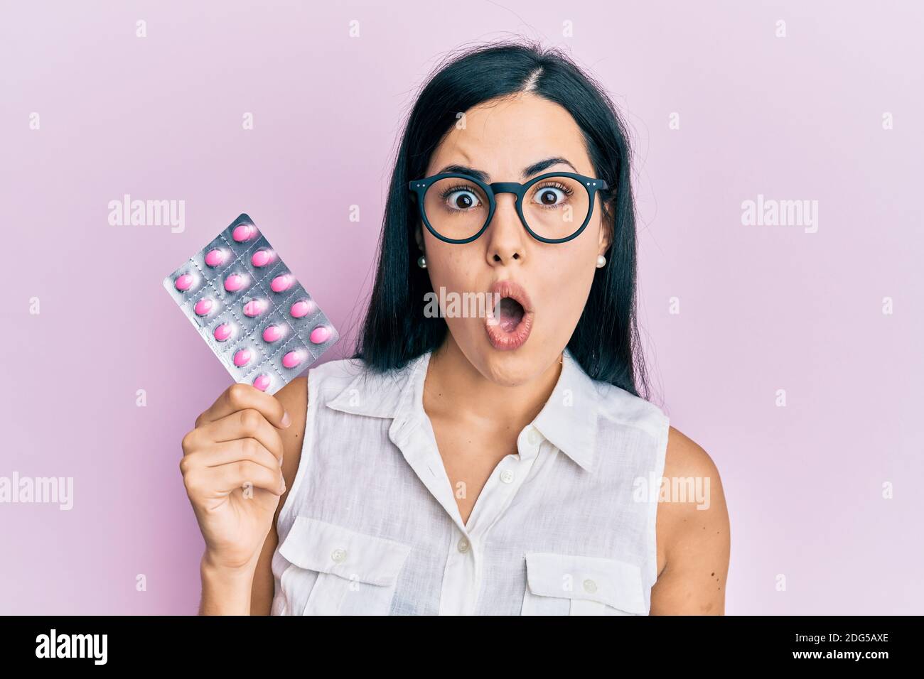 Beautiful young woman holding pills scared and amazed with open mouth