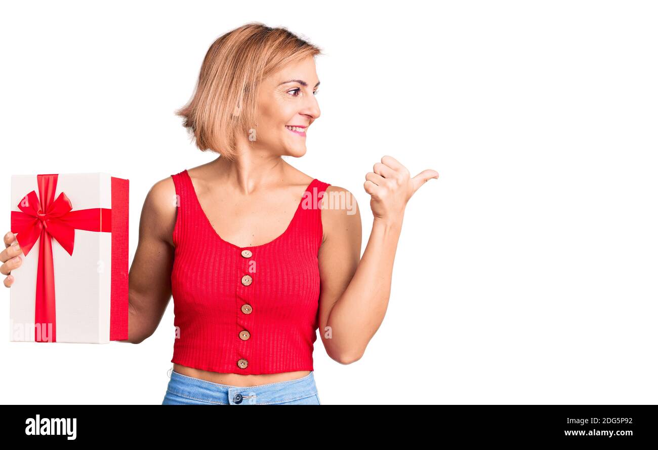 Young blonde woman holding gift pointing thumb up to the side smiling ...