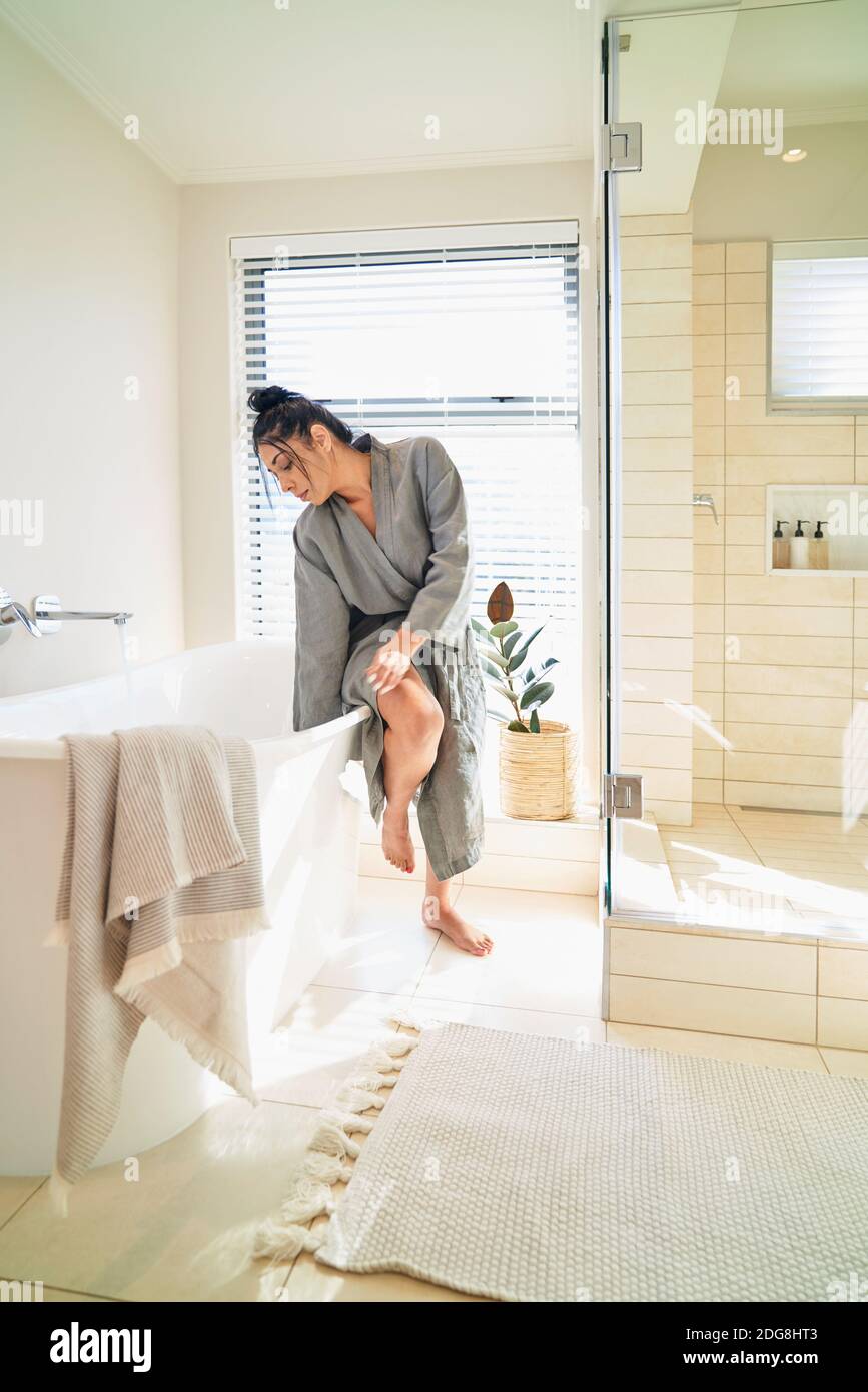 Woman in bathrobe preparing soaking tub for bath in sunny bathroom Stock Photo