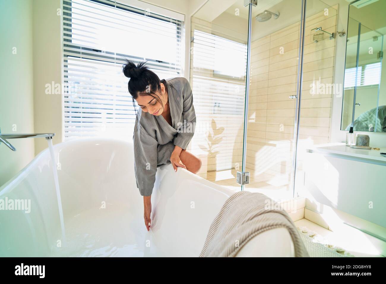 Woman in bathroom preparing soaking tub for bath in sunny bedroom Stock Photo