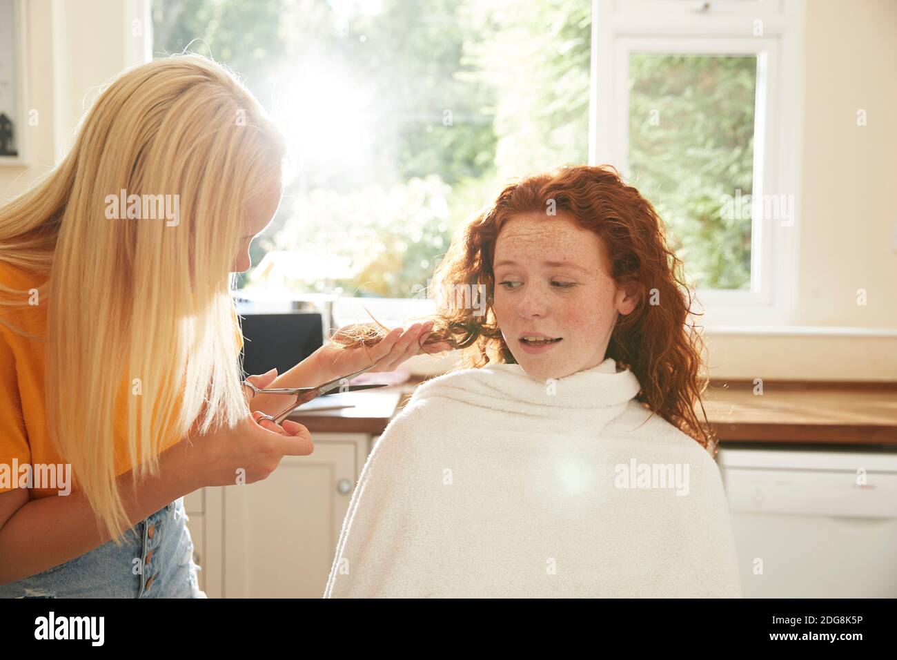 Teenage girl cutting hair for worried friend in sunny kitchen Stock Photo