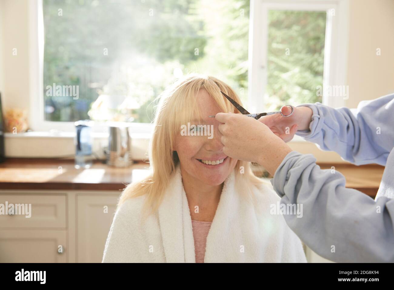 Daughter cutting bangs for happy mother in sunny kitchen Stock Photo