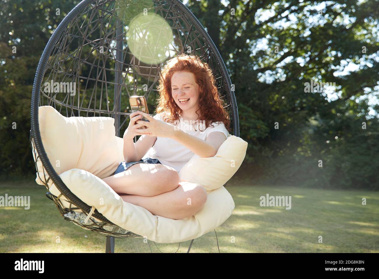 Happy preteen girl using smart phone in swing chair in sunny backyard Stock Photo