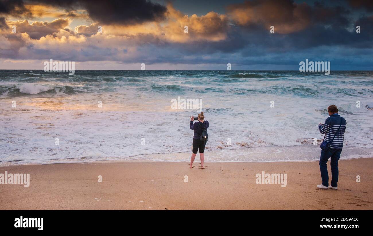 A Panoramic image of a holidaymaker using her mobile phone to photograph a spectacular sunset over Fistral Beach in Newquay in Cornwall. Stock Photo