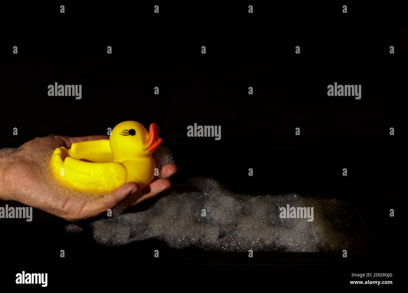 Man's hand holding a yellow rubber duck on a black background with foam Stock Photo