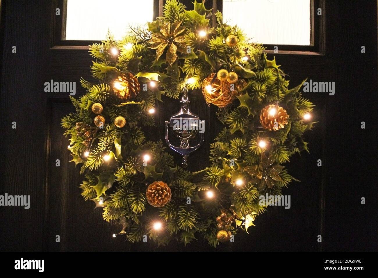 Artificial light-up posh Christmas wreath with gold flowers and pine cones hung on a black door with knocker in the centre in Manchester, England Stock Photo