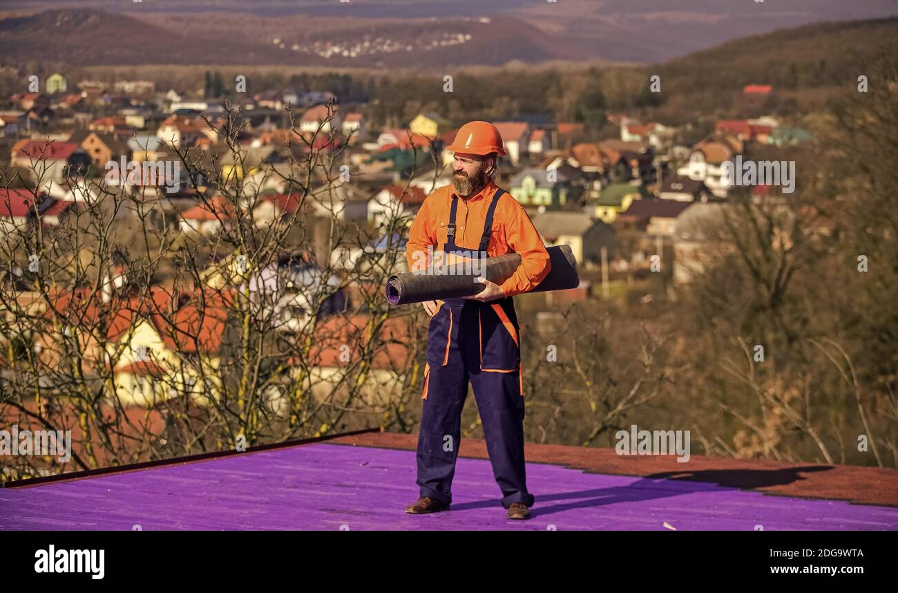 Building house. Apply plastic coatings membranes fiberglass or felt over sloped roofs before applying shingles. Roofer repair roof. Roof installation. Man hard hat work outdoor landscape background. Stock Photo