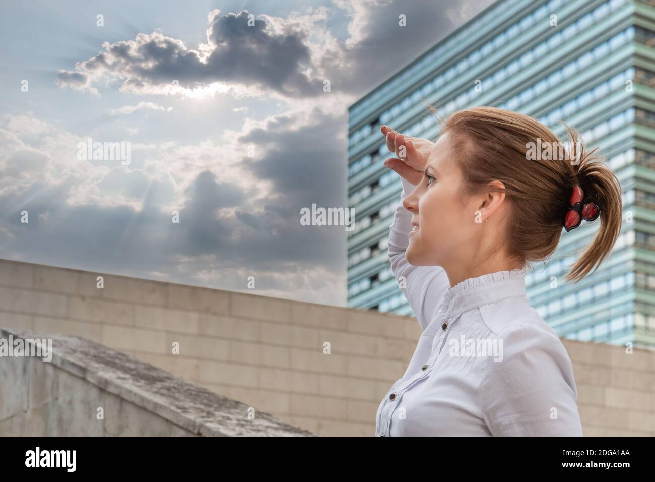 Purposeful woman on urban background and looking ahead at sun between clouds in sky. Businesswoman in cityscape. Successful woman concept. Stock Photo
