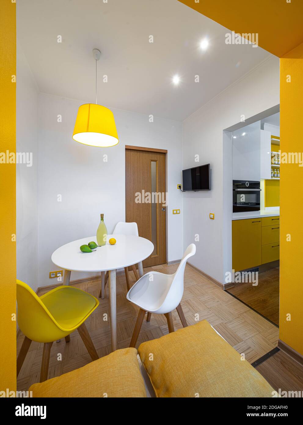 Contemporary interior of kitchen in apartment. Liquor, lemon and pears on table. TV on wall. Wooden door. Yellow and white.  Built-in oven. Stock Photo
