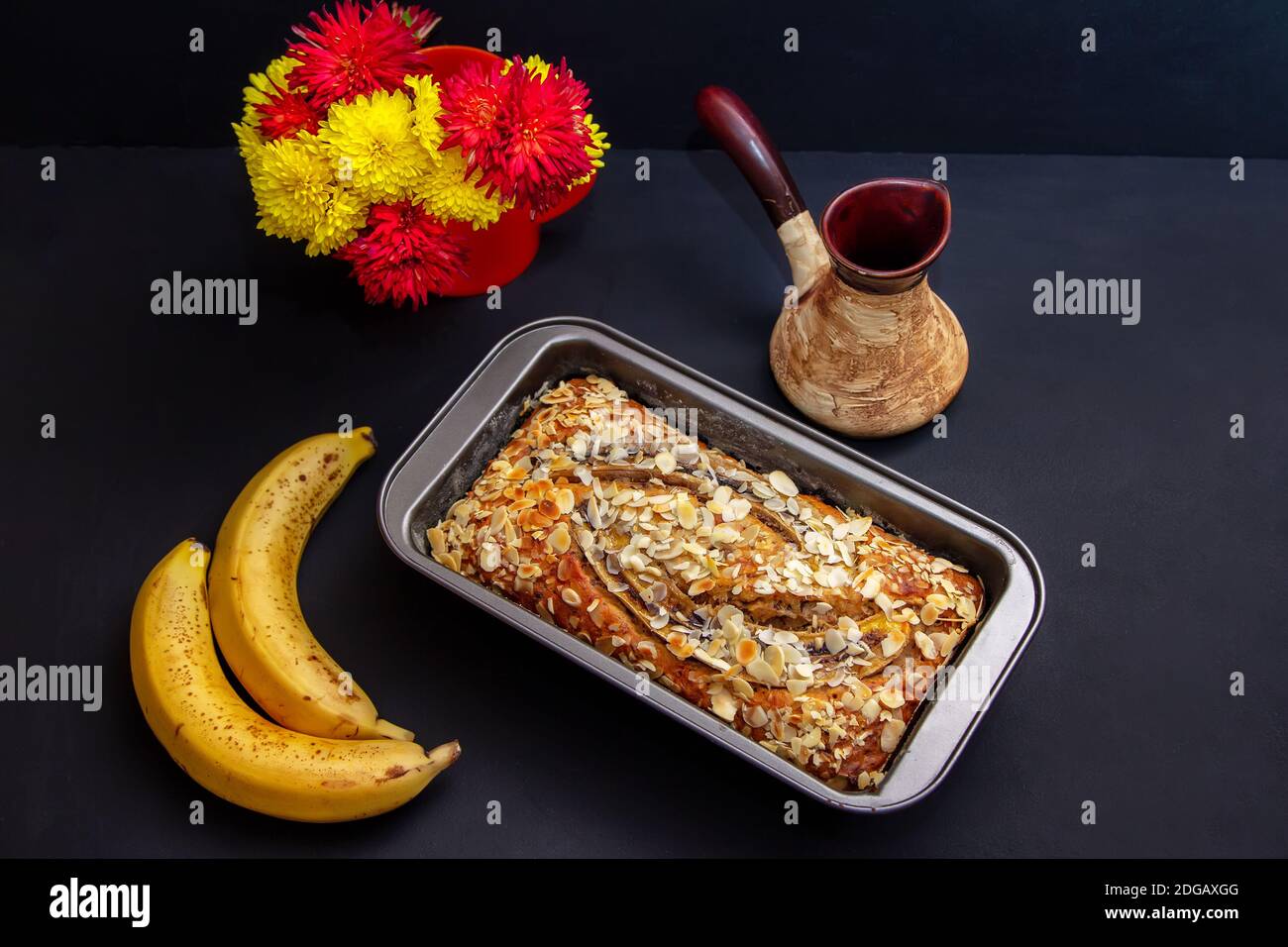 Homemade banana bread with almonds in baking tin, ripe bananas and beautiful colorful flowers on black background Stock Photo