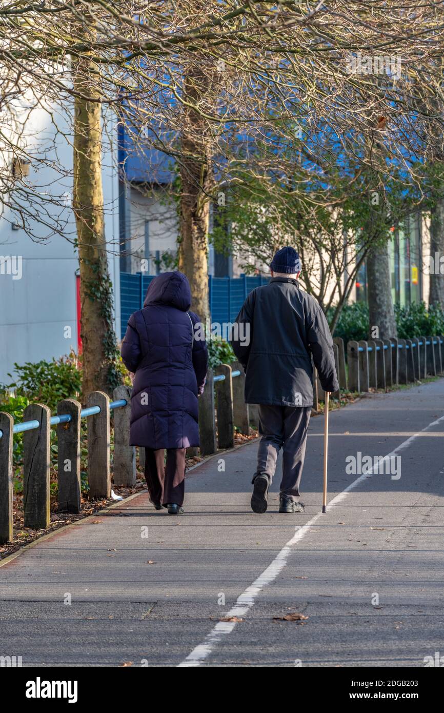 elderly couple walking together along a pathway or sidewalk the man using a walking stick for assistance and aid to walk. Stock Photo