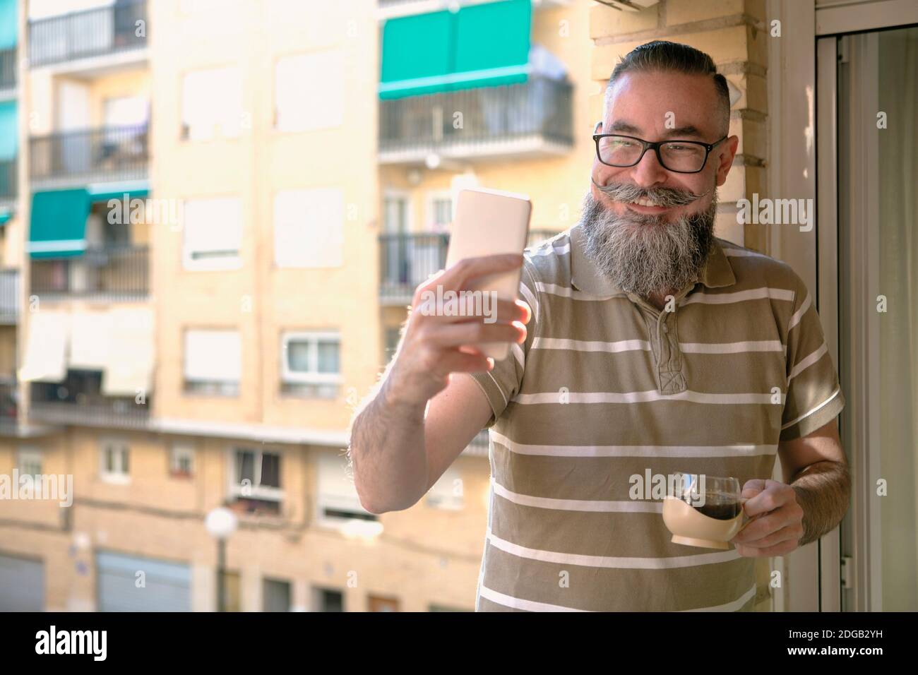 Bearded man with glasses using the cell phone on the balcony of the house to chat and take selfies while smiling and holding a cup of coffee in the ot Stock Photo