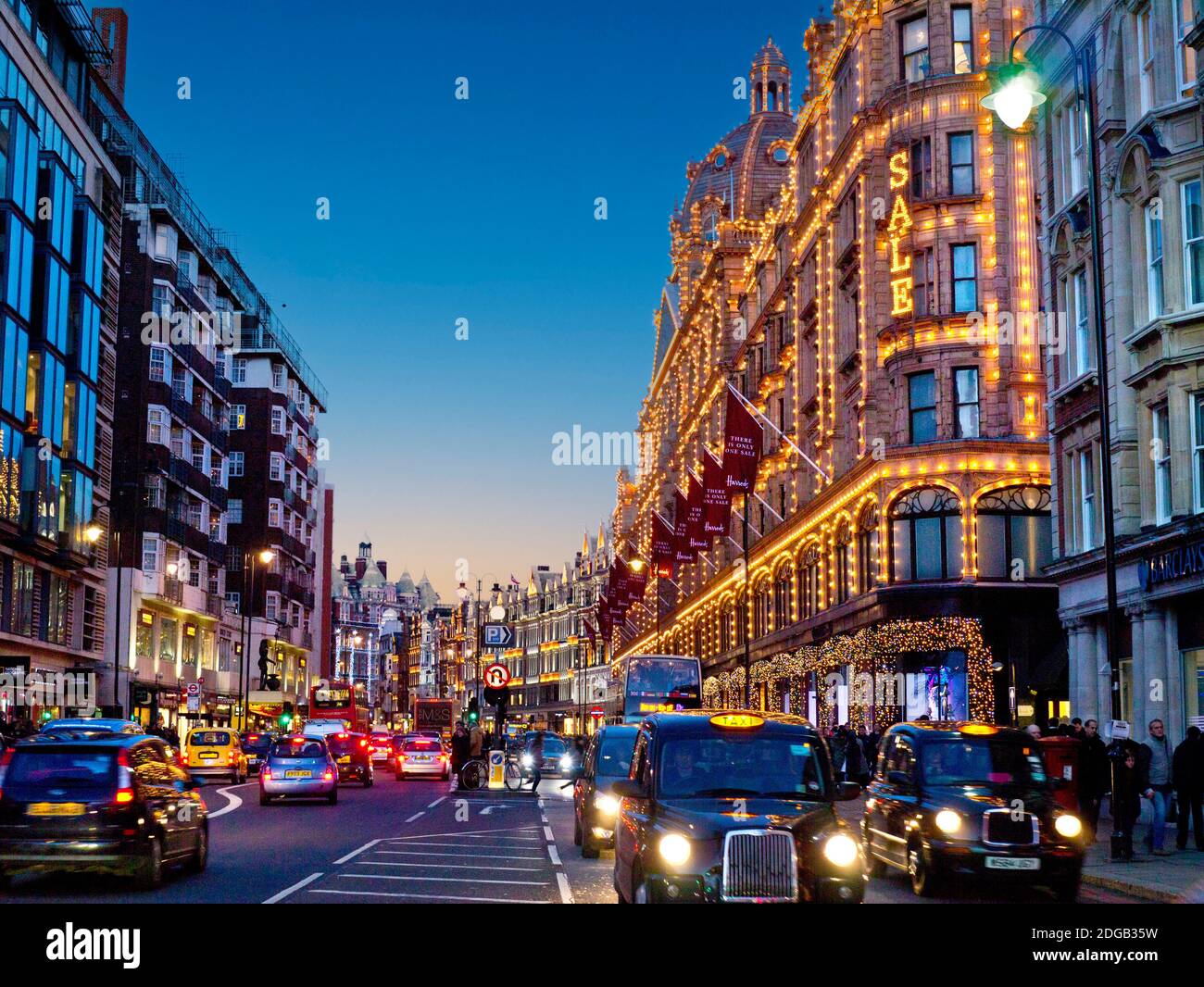 BROMPTON ROAD SUNSET ULEZ HARRODS SALE KNIGHTSBRIDGE SALES TAXIS Brompton Road busy winter sales dusk sunset shops including Harrods store at dusk with lit 'Sale' sign shoppers red buses and black cabs Knightsbridge London SW1 Stock Photo