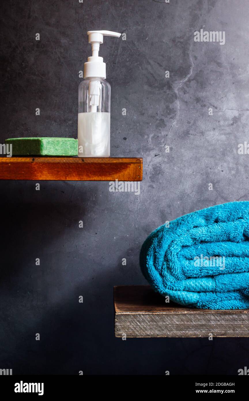 A turquoise towel, a soap dispenser, and a green bar of olive soap on wooden shelves against a gray background Stock Photo