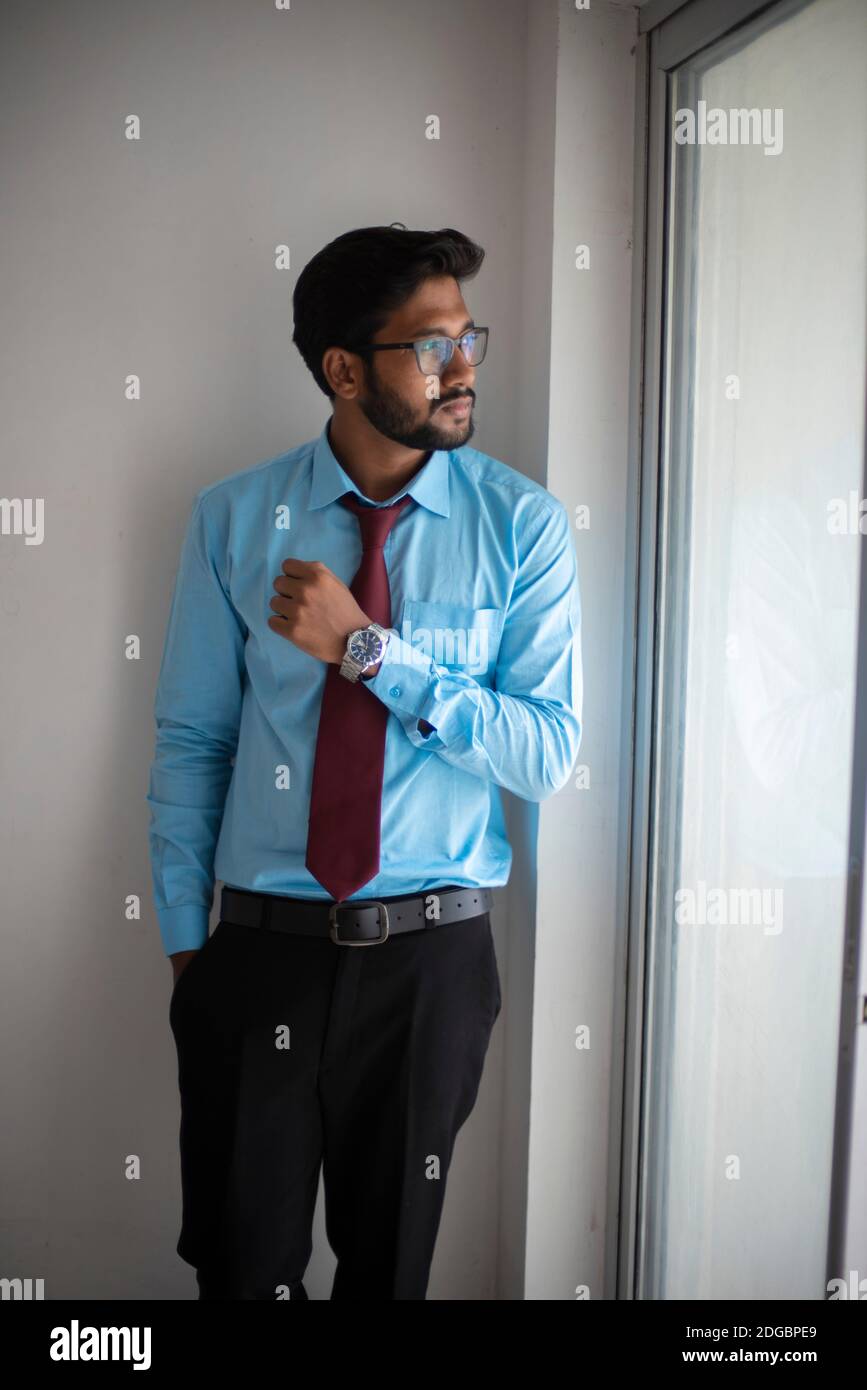 Portrait of an Indian Bengali tall, dark, handsome brunette young man in office wear is standing in front of a glass window in a corporate office/bpo Stock Photo