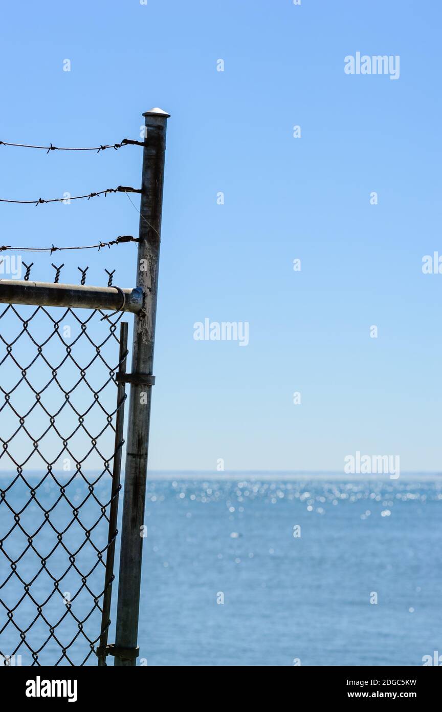 Edge of chain link fence and barbed wires against water and clear blue sky. Stock Photo
