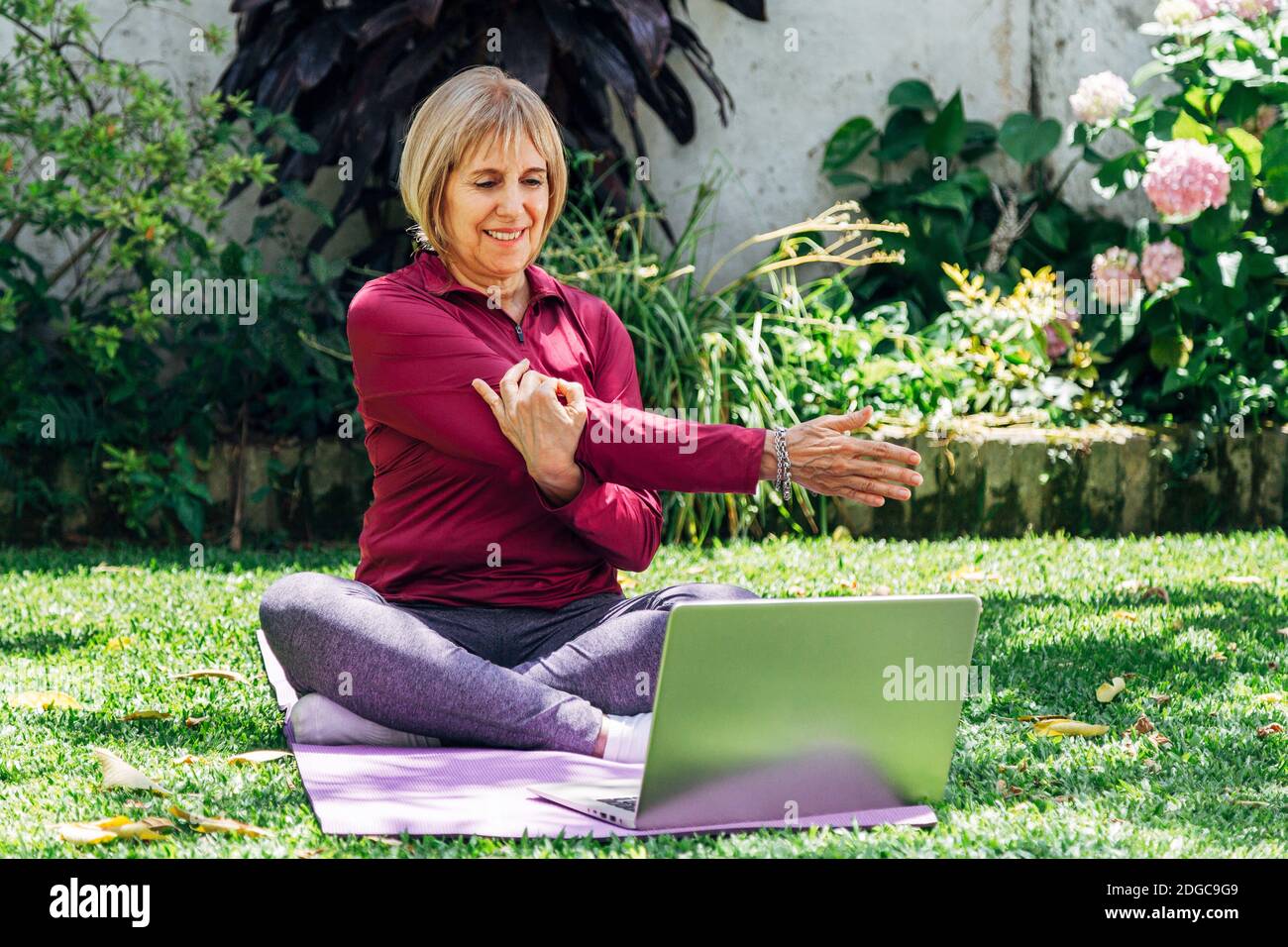 Online sport fitness yoga training workout, senior woman in the garden of her home doing exercises on pilates mat opposite laptop at home. Coach in vi Stock Photo