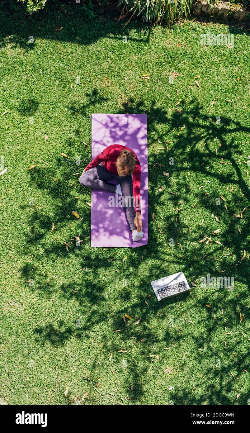 Online sport fitness yoga training workout. Top view of senior woman in the garden of her home doing exercises on pilates mat opposite laptop at home. Stock Photo