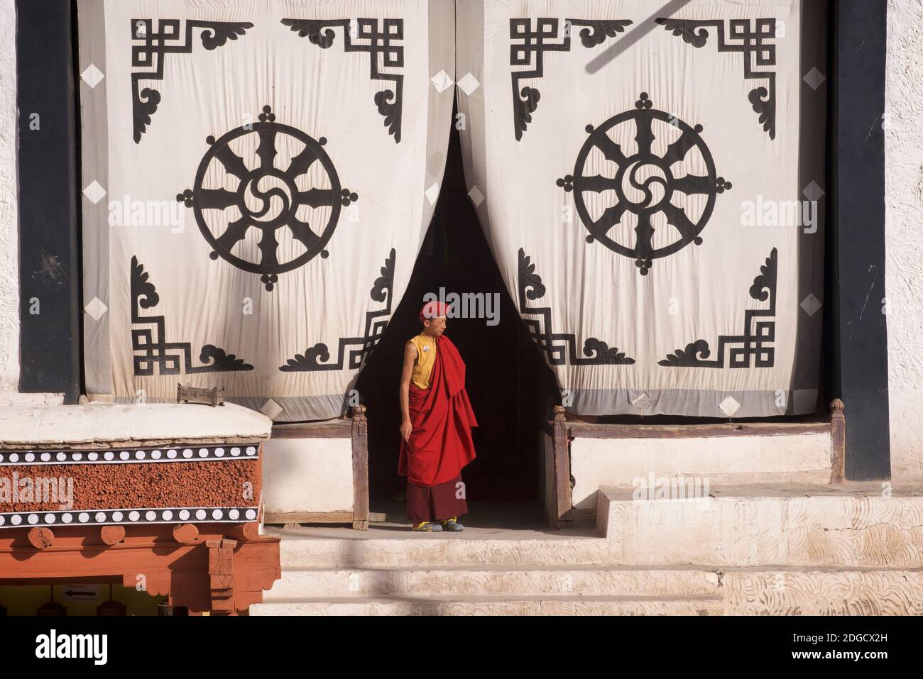 Early morning at Hemis monastery. A young monk  at the entrance to the prayer hall. On the fabric screen at the entrance above is the Dharmachakra - a typical Dharma Wheel with 8 spokes representing the Eightfold Path - The oldest, universal symbol for Buddhism. Hemis monastery, Hemis, Ladakh, Jammu and Kashmir, India. Stock Photo