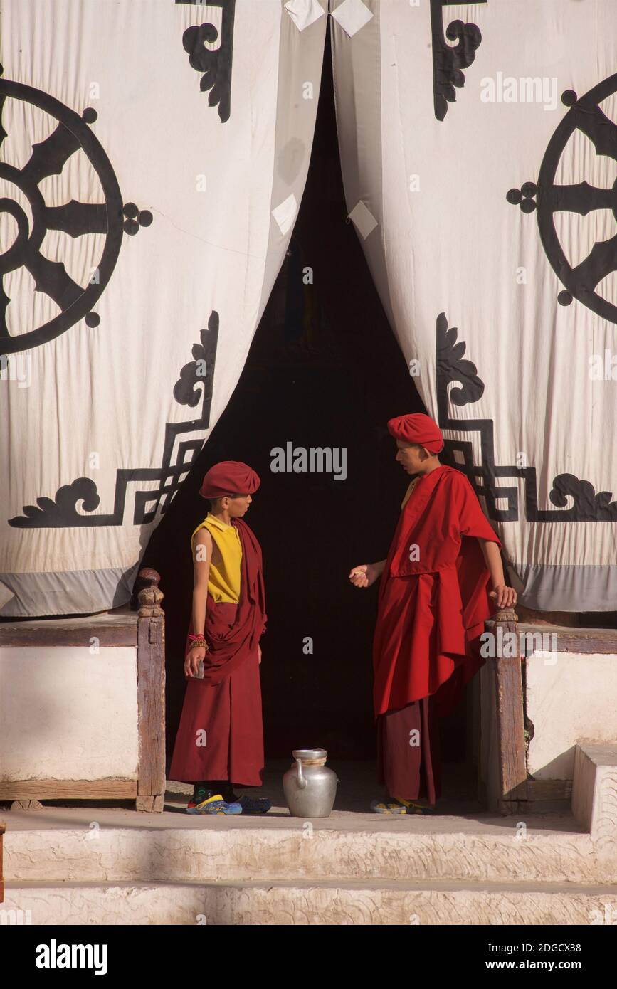 Early morning at Hemis monastery. Two young monks at the entrance to the prayer hall. On the fabric screen at the entrance above is the Dharmachakra - a typical Dharma Wheel with 8 spokes representing the Eightfold Path - The oldest, universal symbol for Buddhism. Hemis monastery, Hemis, Ladakh, Jammu and Kashmir, India. Stock Photo
