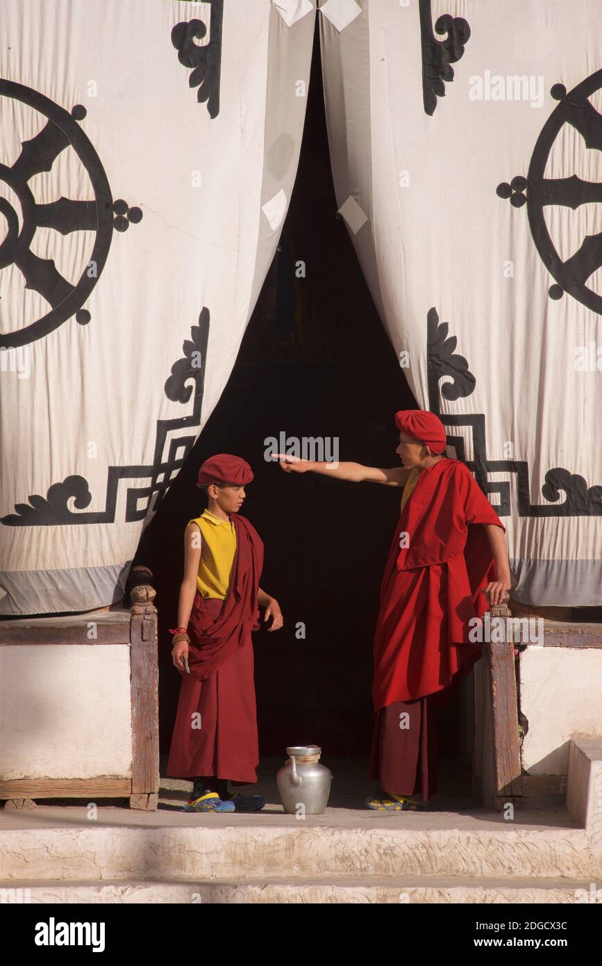 Early morning at Hemis monastery. Two young monks at the entrance to the prayer hall, one directing the other. On the fabric screen at the entrance above is the Dharmachakra - a typical Dharma Wheel with 8 spokes representing the Eightfold Path - The oldest, universal symbol for Buddhism. Hemis monastery, Hemis, Ladakh, Jammu and Kashmir, India. Stock Photo