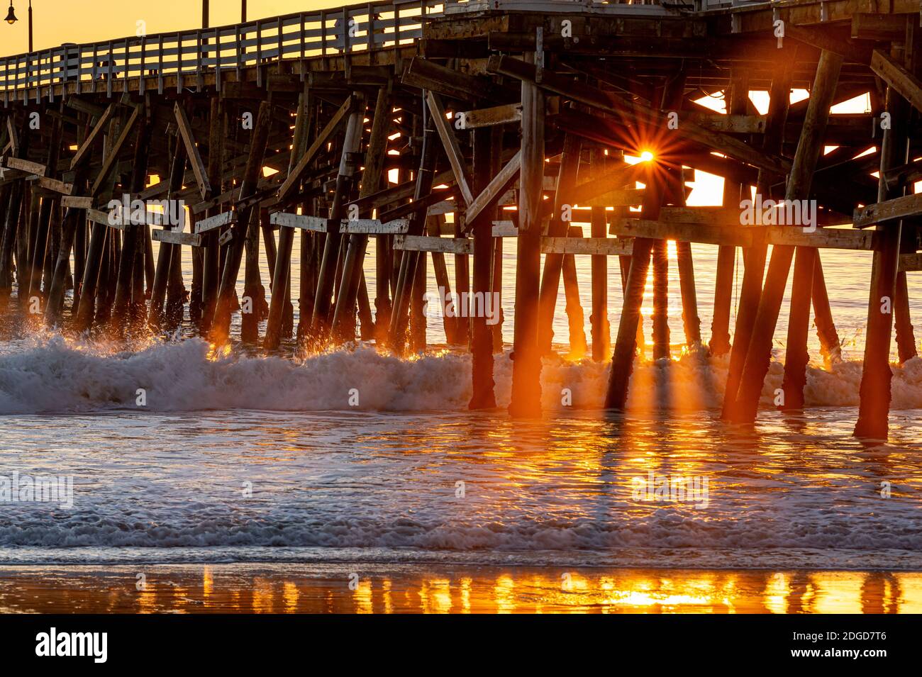 The golden rays of the setting sun stream through a wooden pier structure, shining on the waves and the glassy beach below. Stock Photo