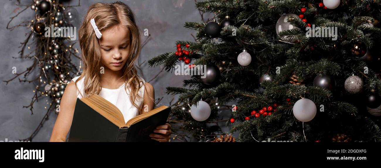 Happy child opening Christmas book. Xmas holiday concept. Girl read a book in decorated living room with Christmas tree. Banner. Copy space. Stock Photo