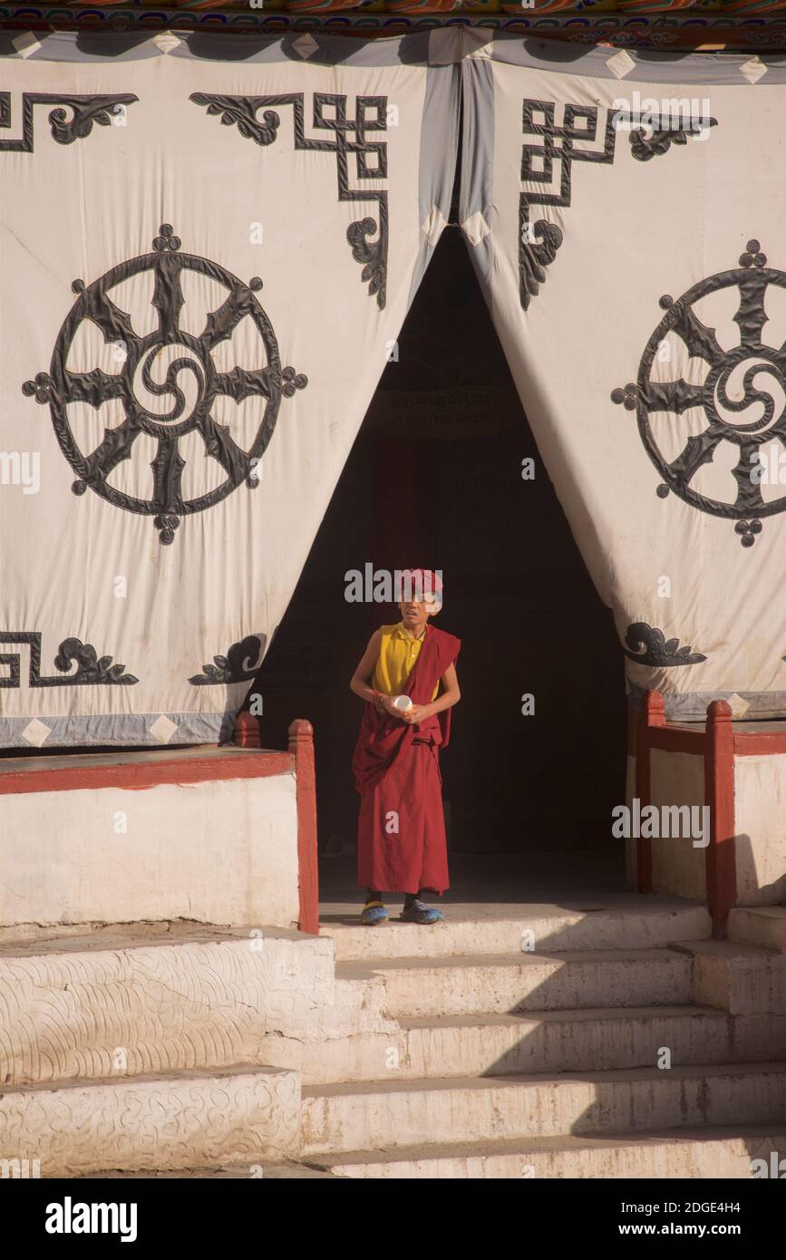 Early morning at Hemis monastery. A young novice monk at the entrance to the prayer hall. On the fabric screen at the entrance above is the Dharmachakra - a typical Dharma Wheel with 8 spokes representing the Eightfold Path - The oldest, universal symbol for Buddhism. Hemis monastery, Hemis, Ladakh, Jammu and Kashmir, India. Stock Photo