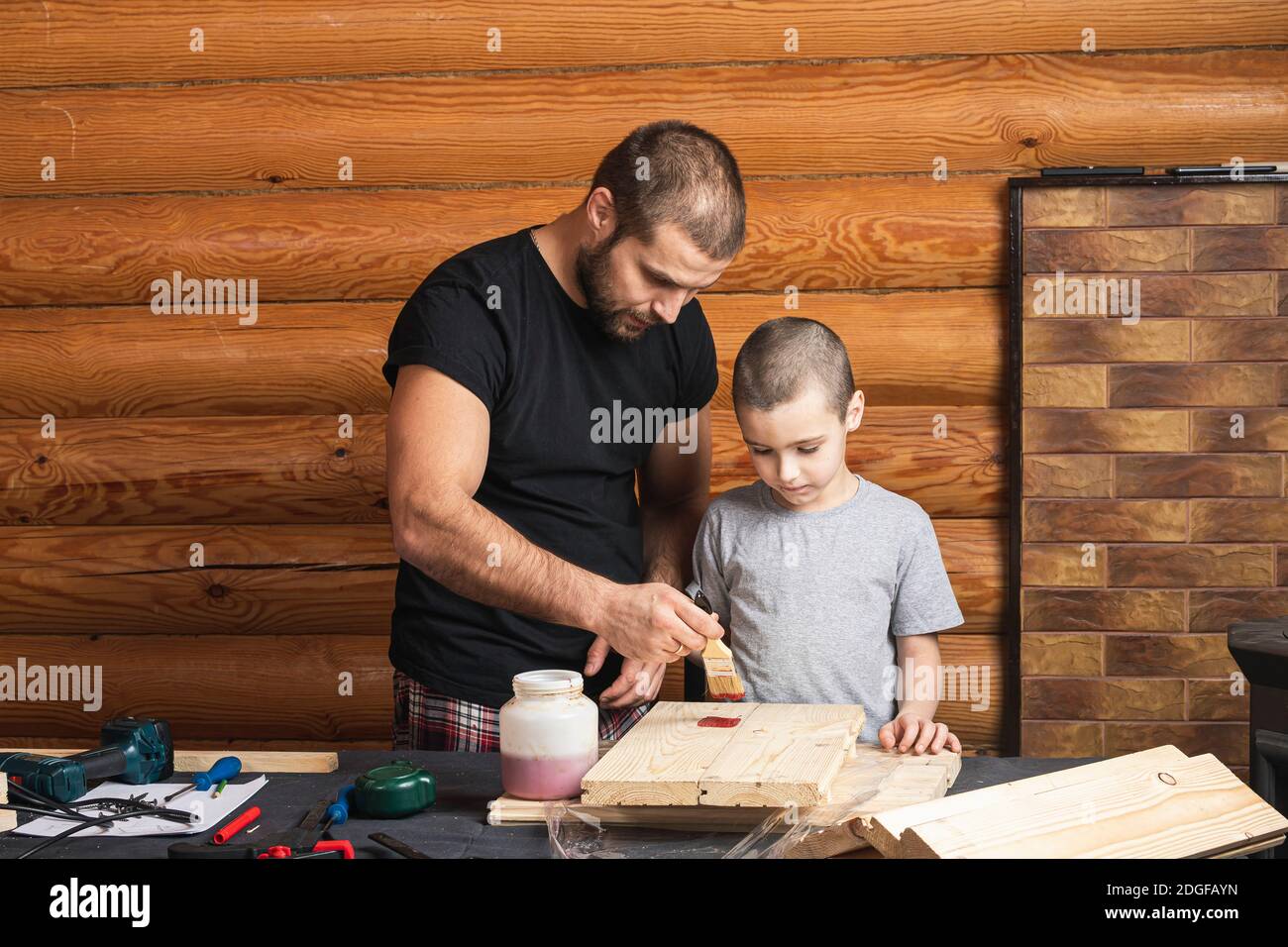 Dad and son are   painting a wooden board with a brush in red,  how to build a bird house, tools and a beam on the table in the workshop. Carpentry tr Stock Photo