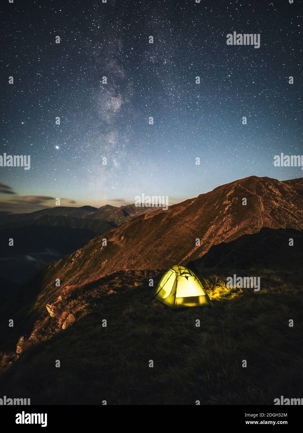 Camping under starry sky and milky way at high altitude on the Carpathian mountains. Illuminated tent in the foreground and majestic mountain peak Stock Photo