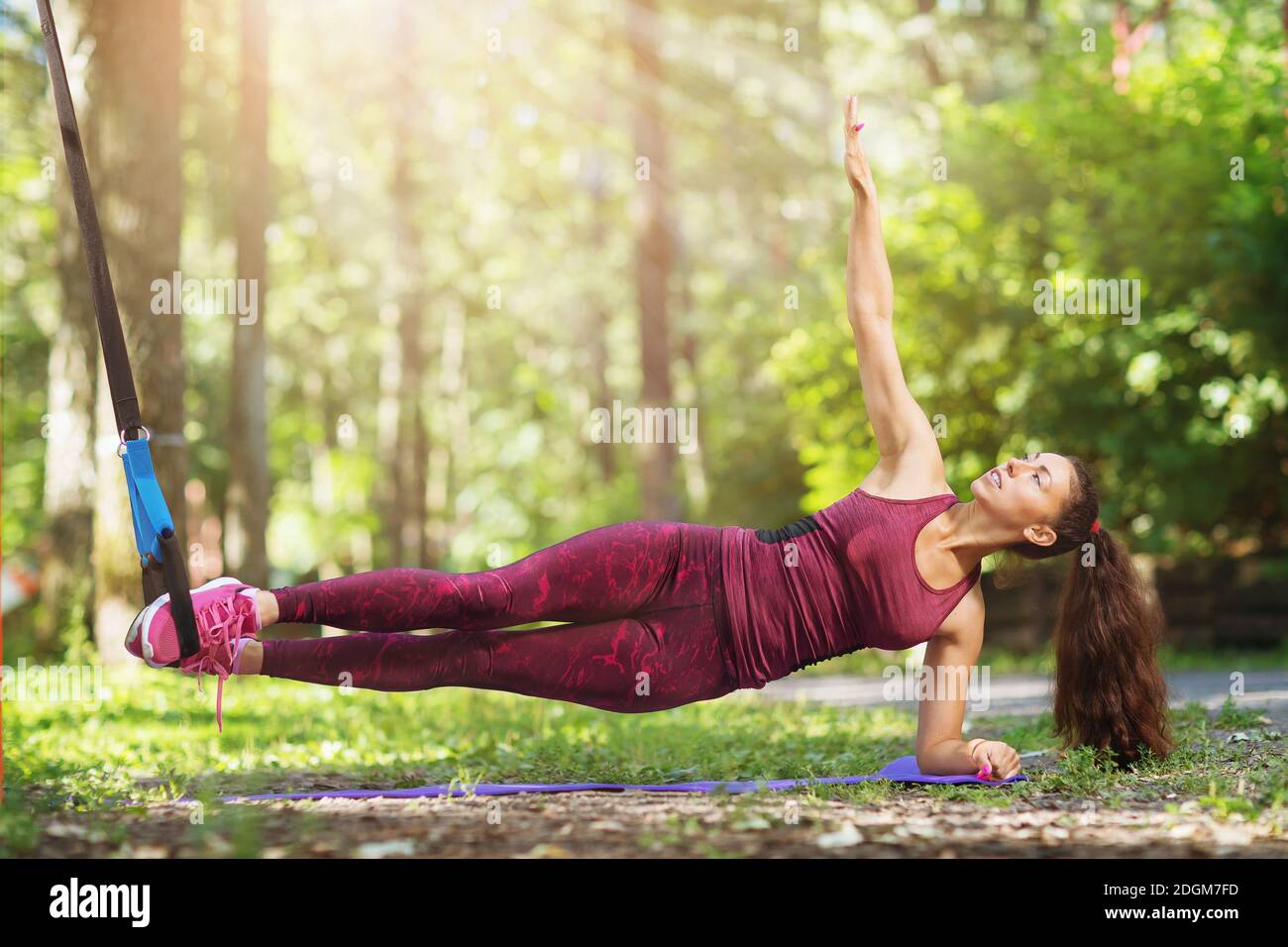 A young woman performs an abdominal exercise on a suspended machine attached to a tree in a park. Stock Photo
